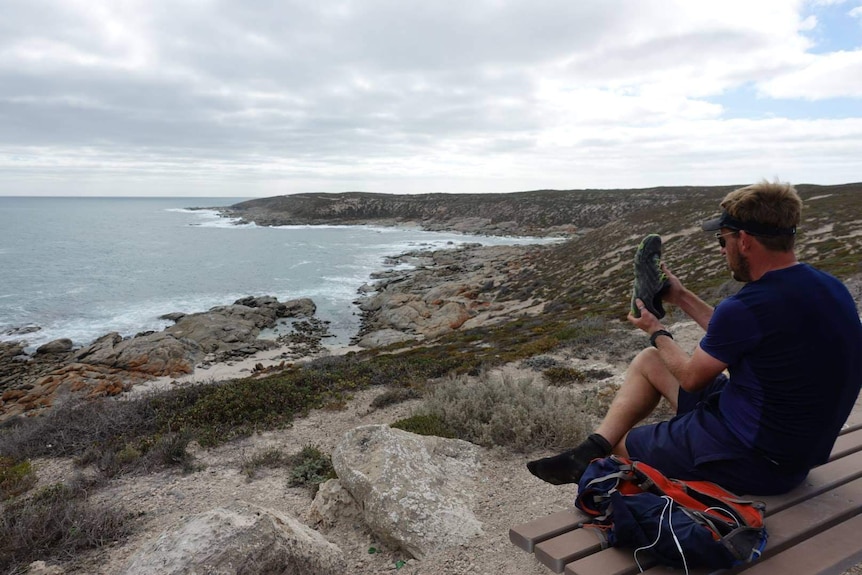 A man changes his shoes on a cliff-top with the ocean stretching along the coast in the background