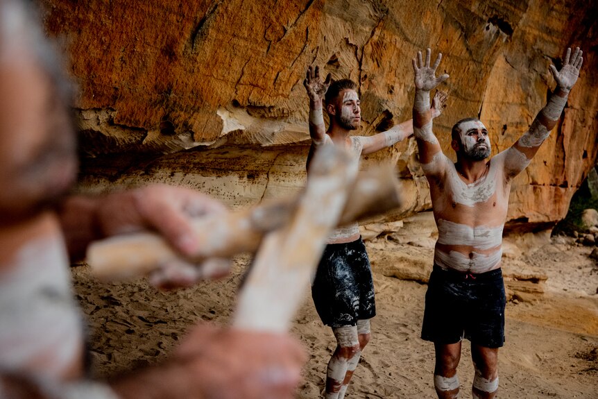 Gomeroi men dance with their arms up, while a man plays rhythm sticks in the foreground.
