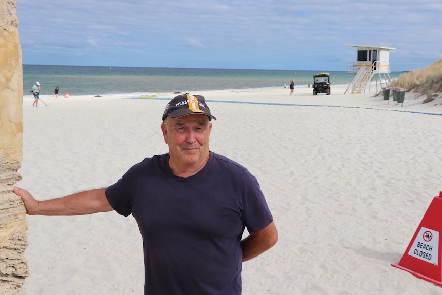 A man in a navy t-shirt and a baseball cap standing on a beach impacted by algae