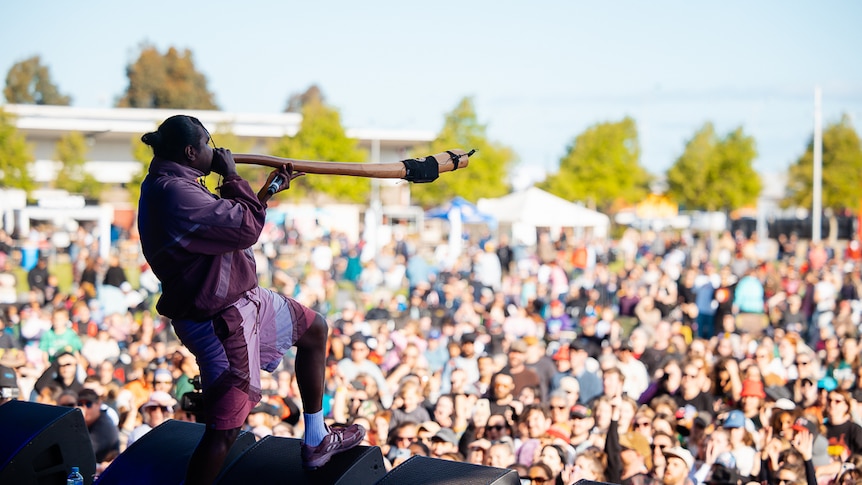 An Indigenous musician in a tracksuit stands with one foot on a wedge and blasts a didgeridoo at a festival crowd.