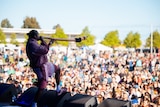 An Indigenous musician in a tracksuit stands with one foot on a wedge and blasts a didgeridoo at a festival crowd.