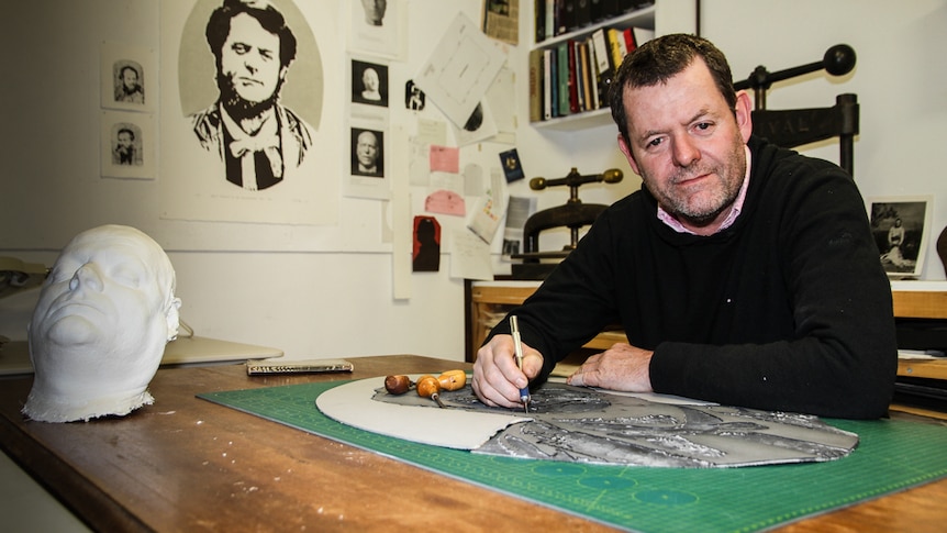 Artist Clayton Tremlett sans beard sitting at his studio desk with his own personal death mask on the desk.