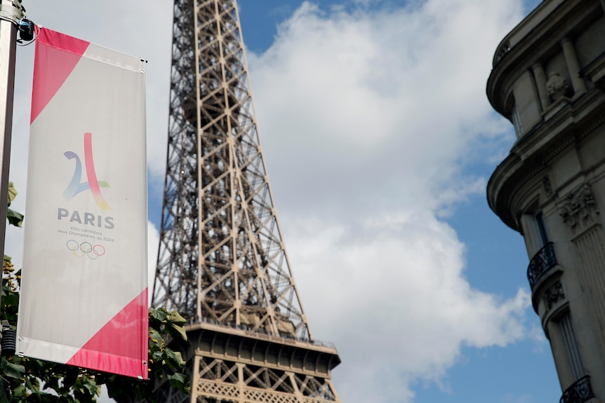A large banner hangs with the logo of the Paris Olympic Games, while next to it stands a large, wrought-iron tower.