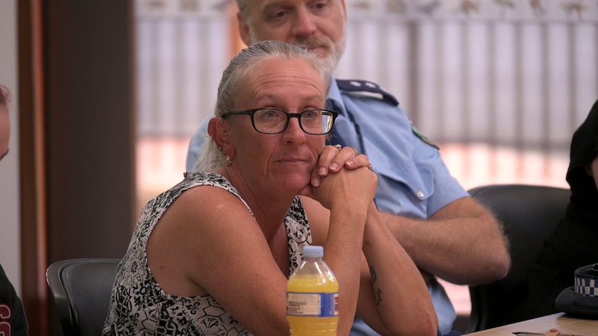 A bespectacled, middle-aged woman with grey hair sits in a meeting room with her hands clasped.