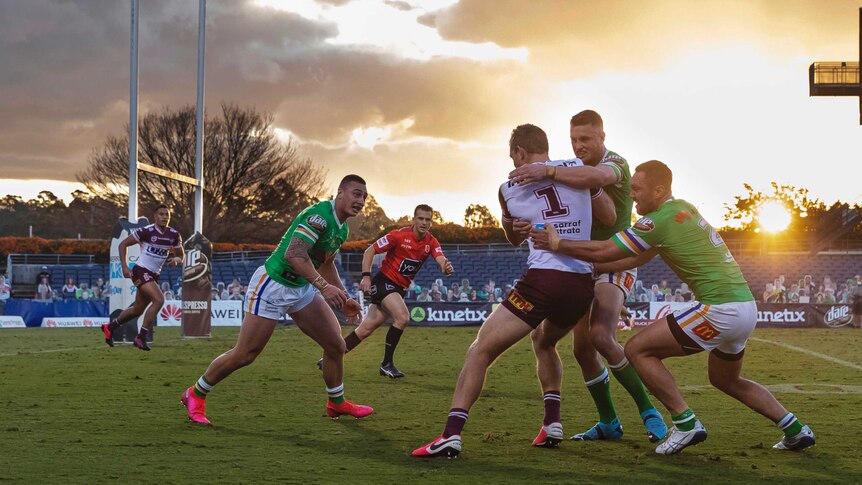 An NRL player is tackled by two defenders near the posts as the sun sets in the background.