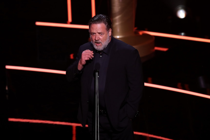 Man with greying beard wears an all-black suit and shirt and speaks into a microphone on a lit stage.