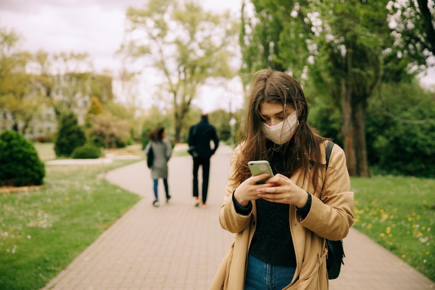 A young woman wearing a face mask looks down at her phone. Two people are visible on the path behind her