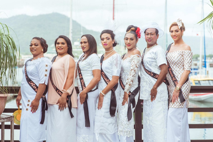 On a jetty, a row of Samoan fa'afafine are shown in bright white formalwear with sashes across them.