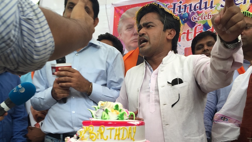Men stand behind a table with a large birthday cake. A poster of Donald Trump is in the background.