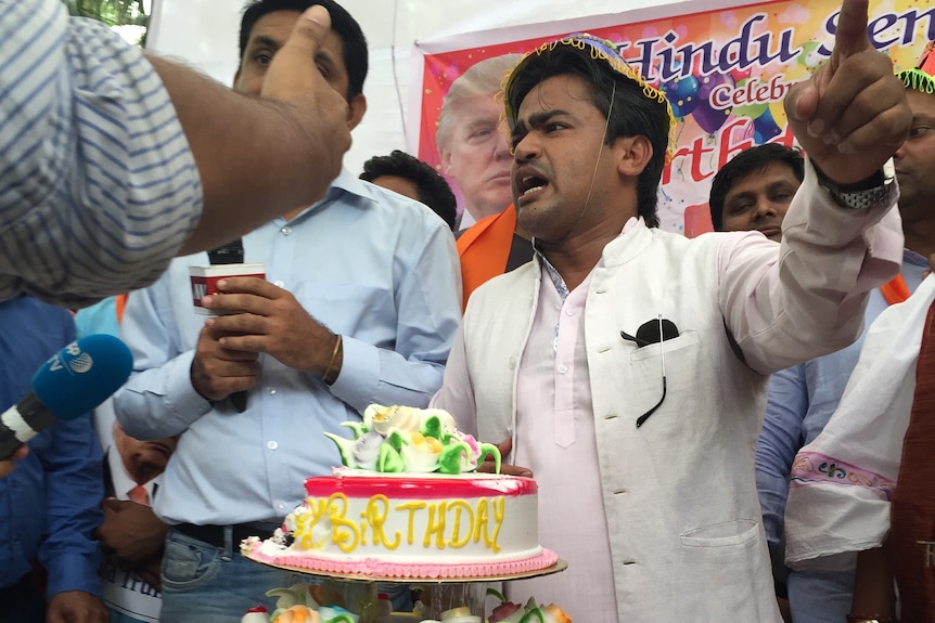 Men stand behind a table with a large birthday cake. A poster of Donald Trump is in the background.