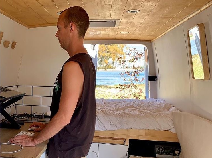 Man standing at a laptop inside the back of a van. The back doors to the van open out to a water view.