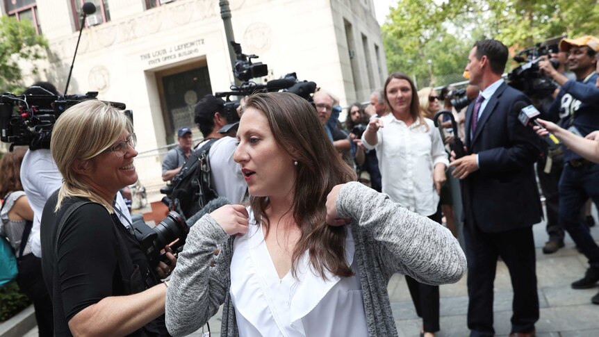 A woman in a white blouse and grey cardigan walks through a media scrum outside a courthouse.
