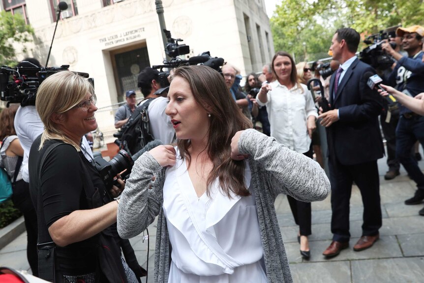 A woman in a white blouse and grey cardigan walks through a media scrum outside a courthouse.