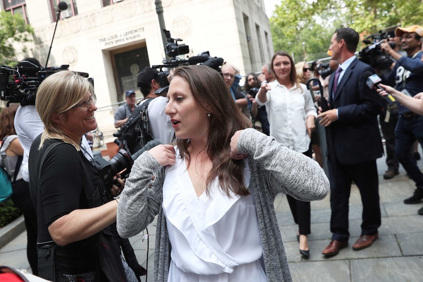 A woman in a white blouse and grey cardigan walks through a media scrum outside a courthouse.