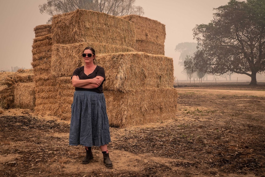Sarah Kippel wears a blue skirt, black t-shirt and sunglass. She stands in front of pile of donated hay, thick smoke haze.