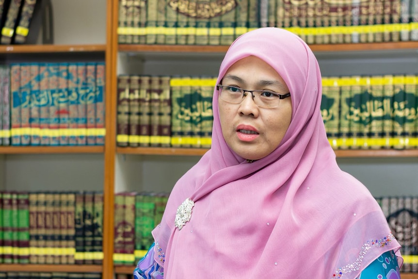 A woman wearing a pink hijab talks to someone off-camera. Bound books are in the background.