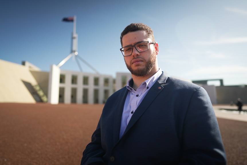 A young white man with short brown hair and glasses out the front of Parliament House in Canberra. He is a wheelchair user
