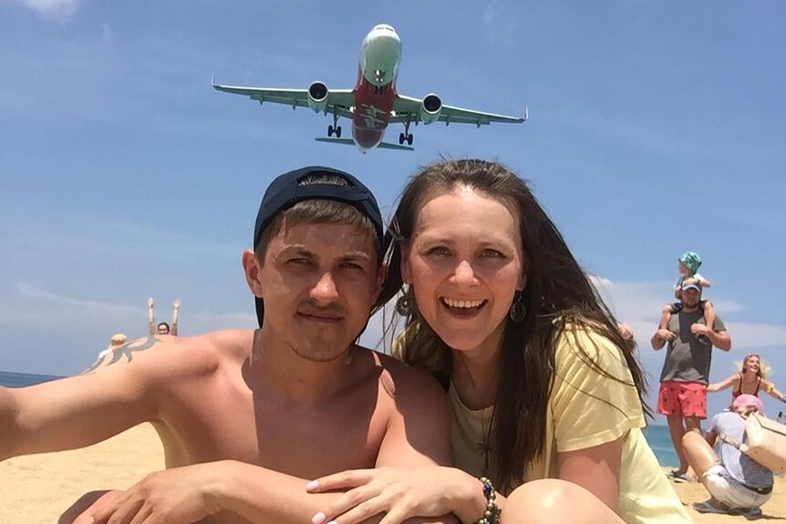 A young couple crouch together on the sand at the beach as a plane flies above their heads