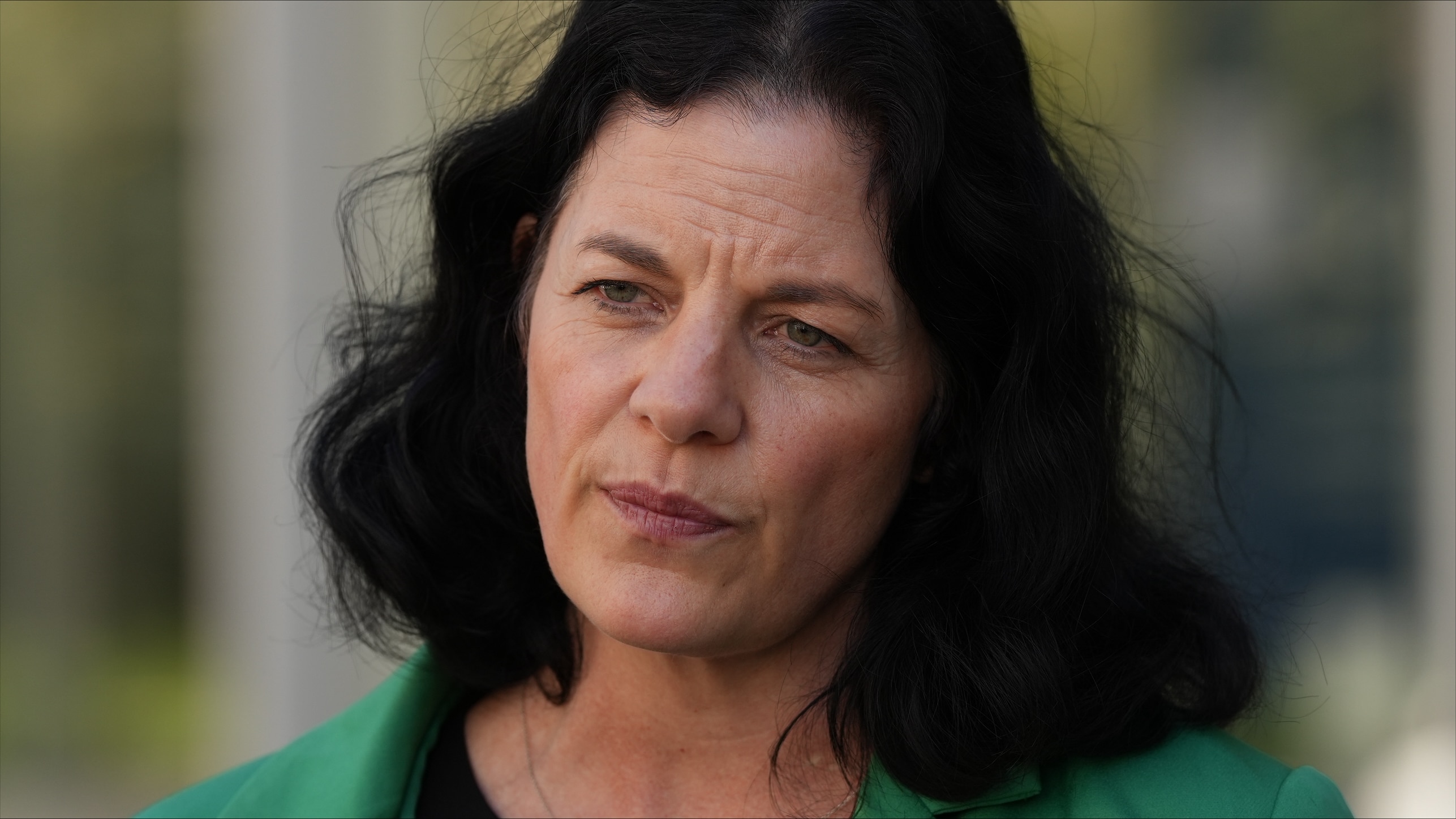 A woman with black shoulder-length wavey hair in a black top and green blazer speaks to media.