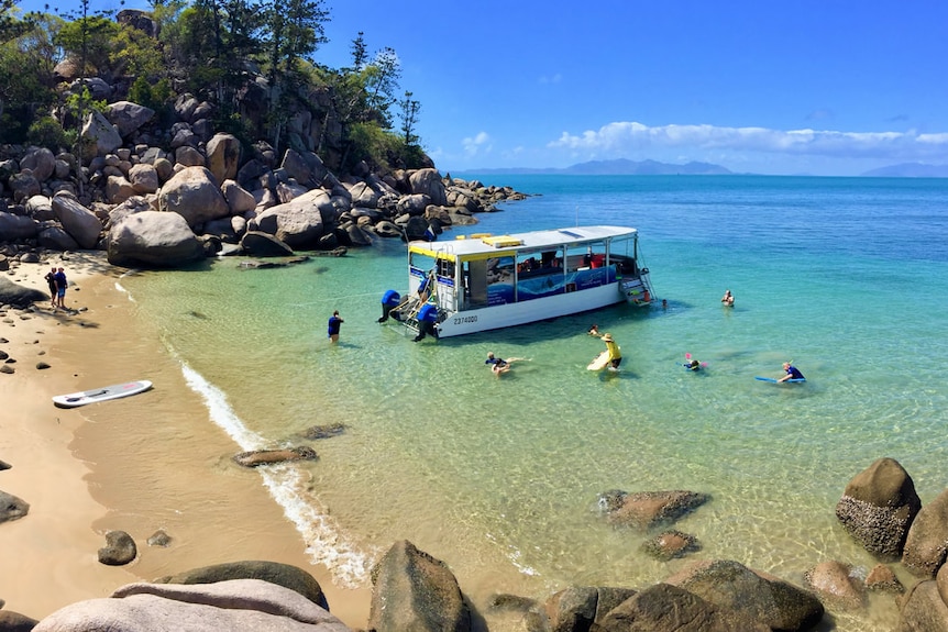 A birds eye view picture of a small boat in a secluded beach on Magnetic island, swimmers float around the boat in clear water.