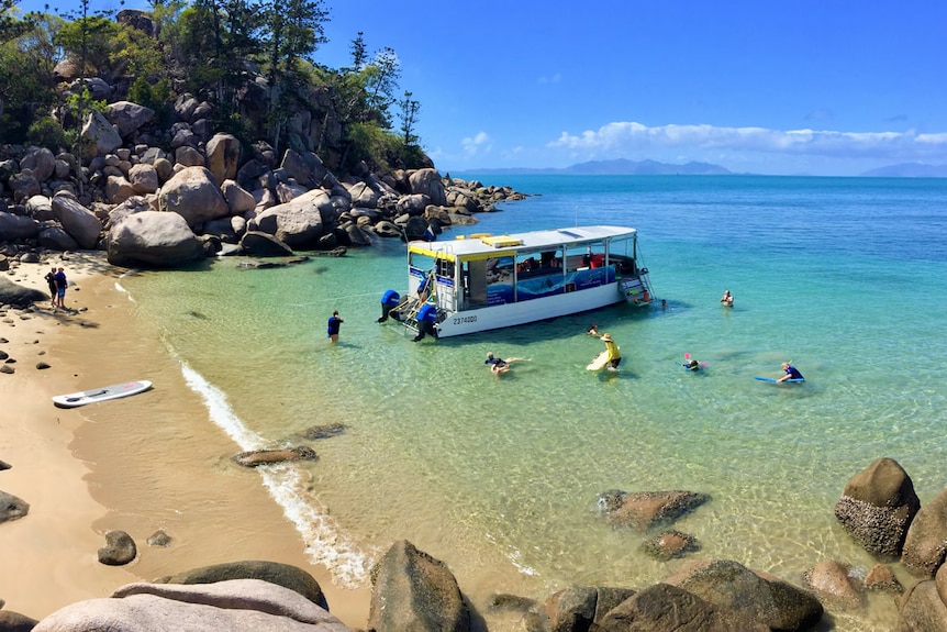 Una imagen a vista de pájaro de un pequeño bote en una playa aislada en la isla Magnetic, los nadadores flotan alrededor del bote en agua clara.