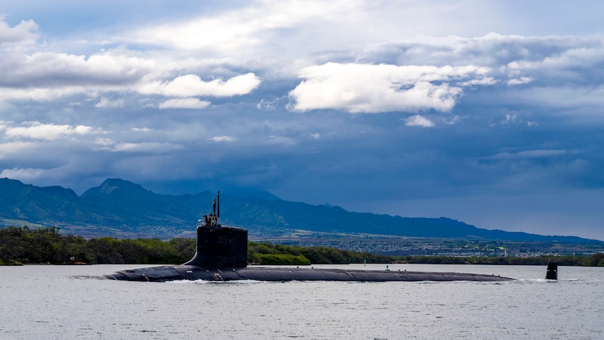 A submarine is seen on the surface of the water in front of mountains and clear skies.