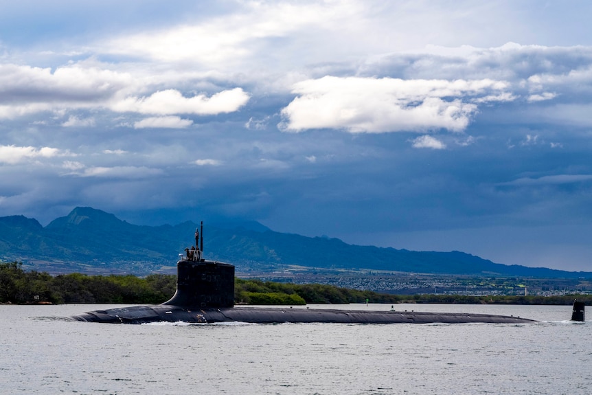 A submarine is seen on the surface of the water in front of mountains and clear skies.