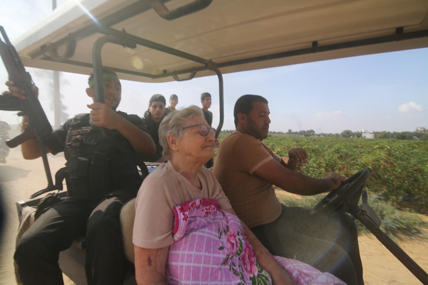 An elderly woman sits in the front seat of a golf cart driven by Palestinian militants, one holding a gun