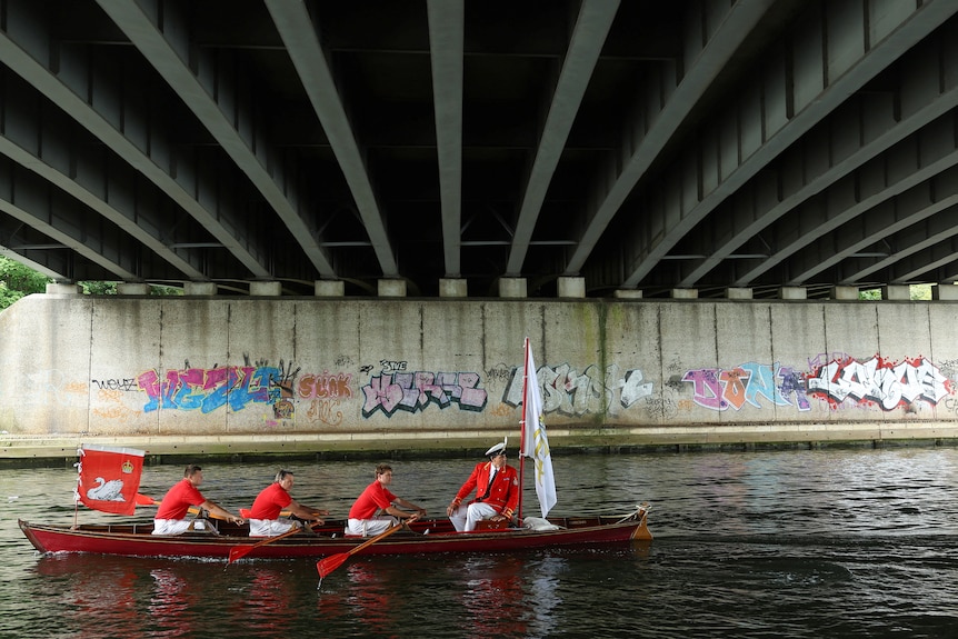 Four men dressed in red and white row a boat under a bridge