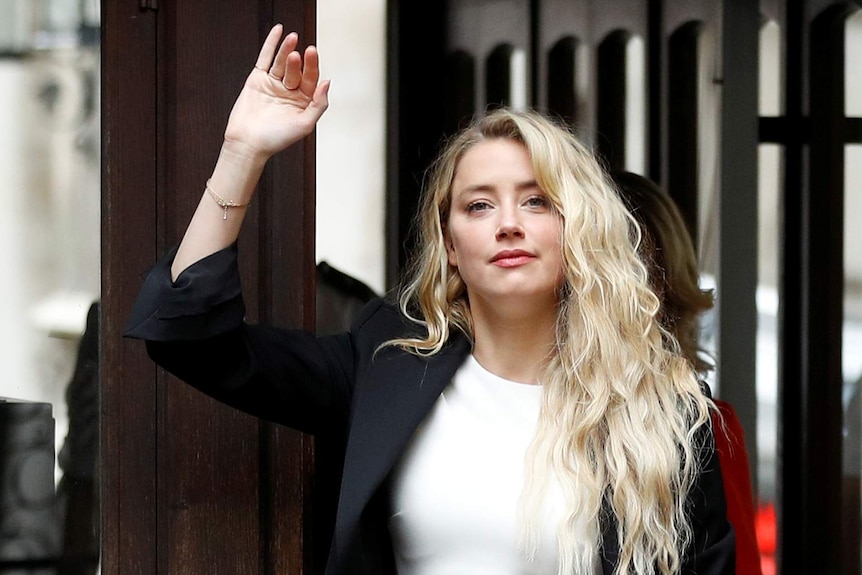 A woman with blond hair waves to people, she is wearing a black jacket and white top underneath.