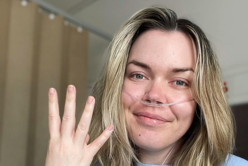 A woman with long blonde hair smiles at the camera in a hospital bed, holding up 4 fingers, to signify how many surgeries.