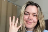 A woman with long blonde hair smiles at the camera in a hospital bed, holding up 4 fingers, to signify how many surgeries.
