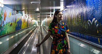 Writer Vanessa Berry stands on an escalator staring up at the camera, wearing a brightly printed day dress.