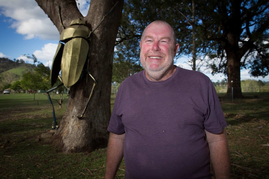 A smiling man pictured outside in front of a tree that has a large golden cicada sculpture attached to it.