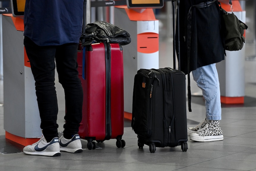Legs of two passengers are seen standing beside their luggage while waiting at an airport
