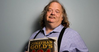 Food critic Jonathan Gold wearing blue shirt and braces and holding a copy of LA Times' 101 Restaurant's edition.