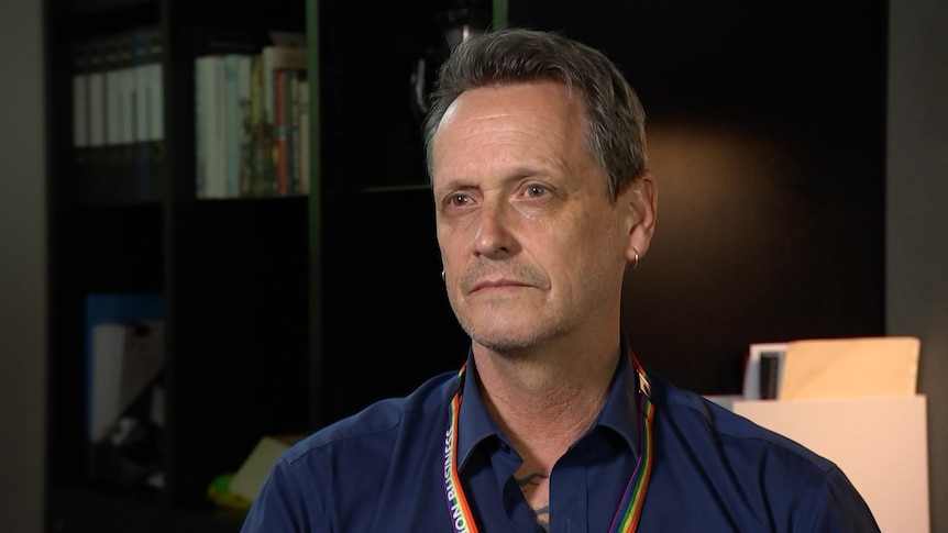 Andy Meddick sits in an office space wearing a navy shirt and a rainbow lanyard.