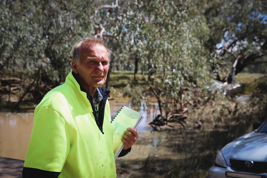 Gary Langley in yellow high vis jumper stands in front of Graddle Creek with car in right corner, holding paperwork