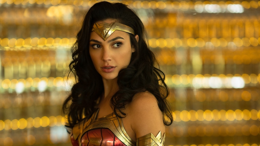 Gal Gadot in Wonder Woman costume and hair out, with sparkling backdrop blurred in background.
