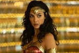 Gal Gadot in Wonder Woman costume and hair out, with sparkling backdrop blurred in background.