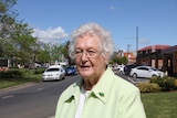 An elderly, bespectacled woman stands near a street in a country town.