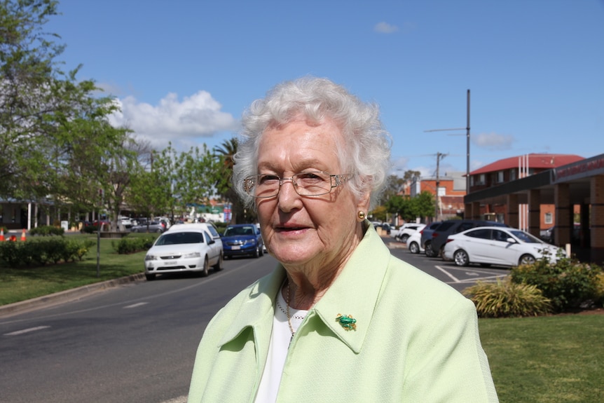 An elderly woman in a green jacket looking at the camera while standing in a country town.