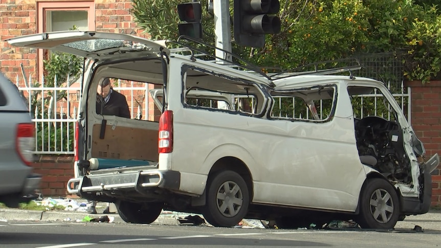 A crumpled white van sits on a residential street in daylight, with police officers walking around it.