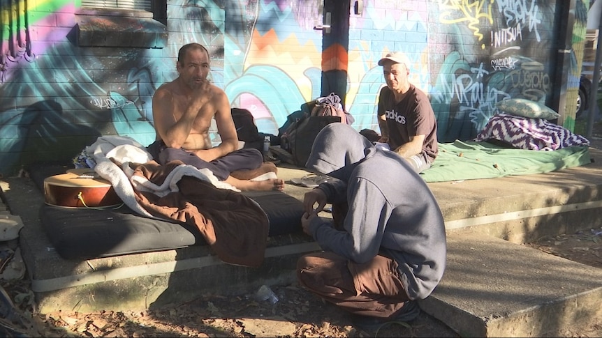 Three homeless men sit on the street among their possessions.