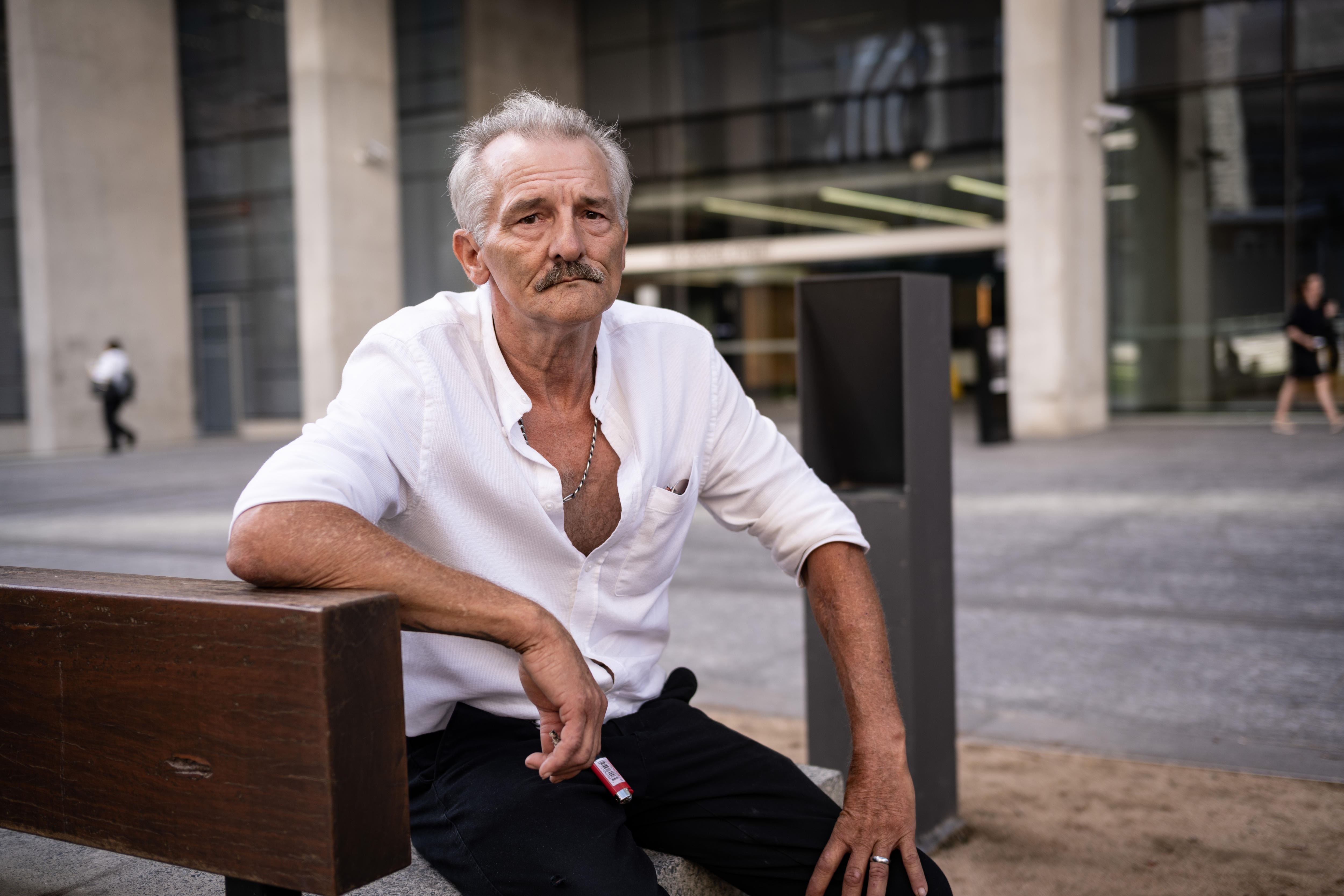 Un hombre mayor con camisa abotonada y pantalones oscuros está sentado frente a un gran tribunal.