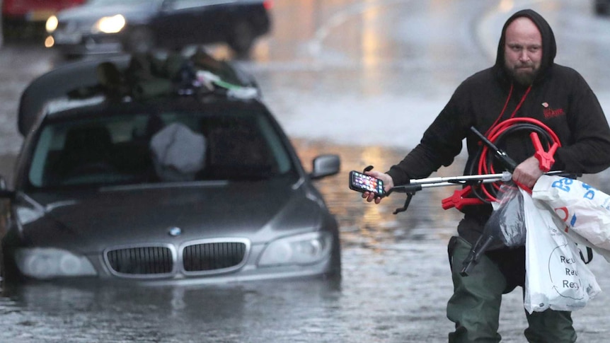 A man wearing a black jumper wades through ankle-deep flood waters after gathering items from his BMW car submerged in floods.