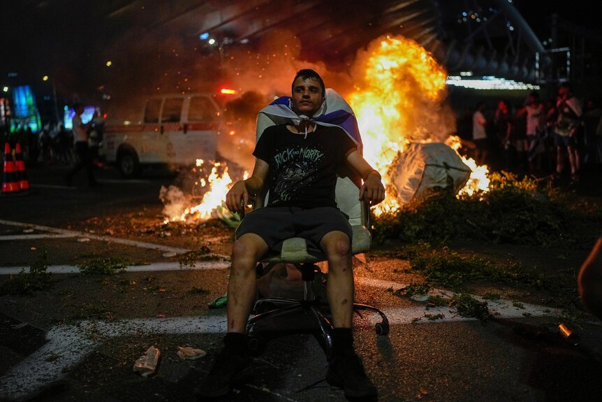 A man sits in a ruined chair on the street while a car blazes behind him 