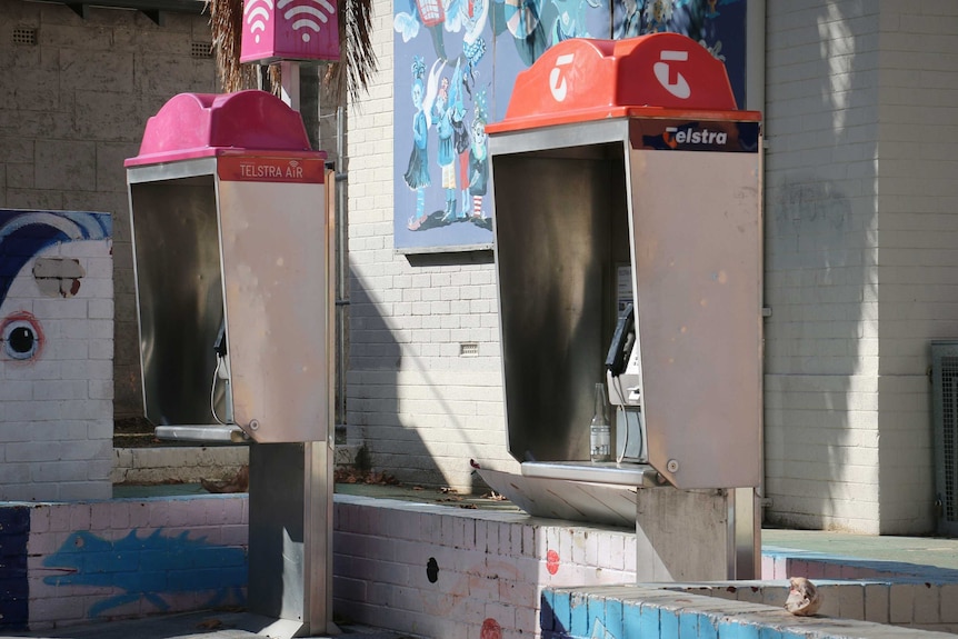 Two aging public phone boxes positioned on a sidewalk, with an empty beer bottle left inside one.