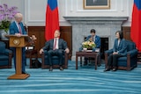 An Australian MP stands at a lecturn with three people, including Taiwan's President on stage behind him. 