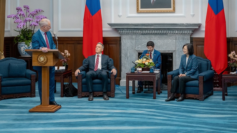 An Australian MP stands at a lecturn with three people, including Taiwan's President on stage behind him. 